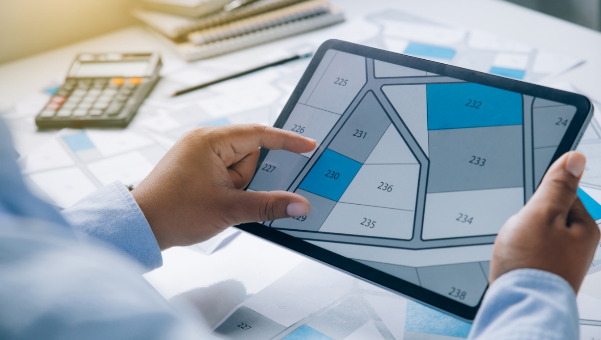 A person holds a tablet displaying a property plot map with numbered sections, focusing on plot 230. A calculator, pen, and additional documents are visible in the background.