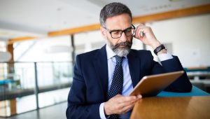 A man in a blue business suit wearing glasses looks at a tablet while standing at a wood countertop in an open concept space.