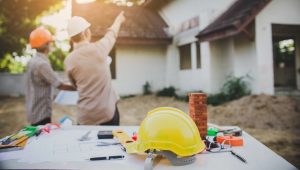 Two construction workers wearing hard hats discuss plans in front of a house under construction, with one pointing towards the building. A table in the foreground holds a yellow hard hat, blueprints, and various tools.