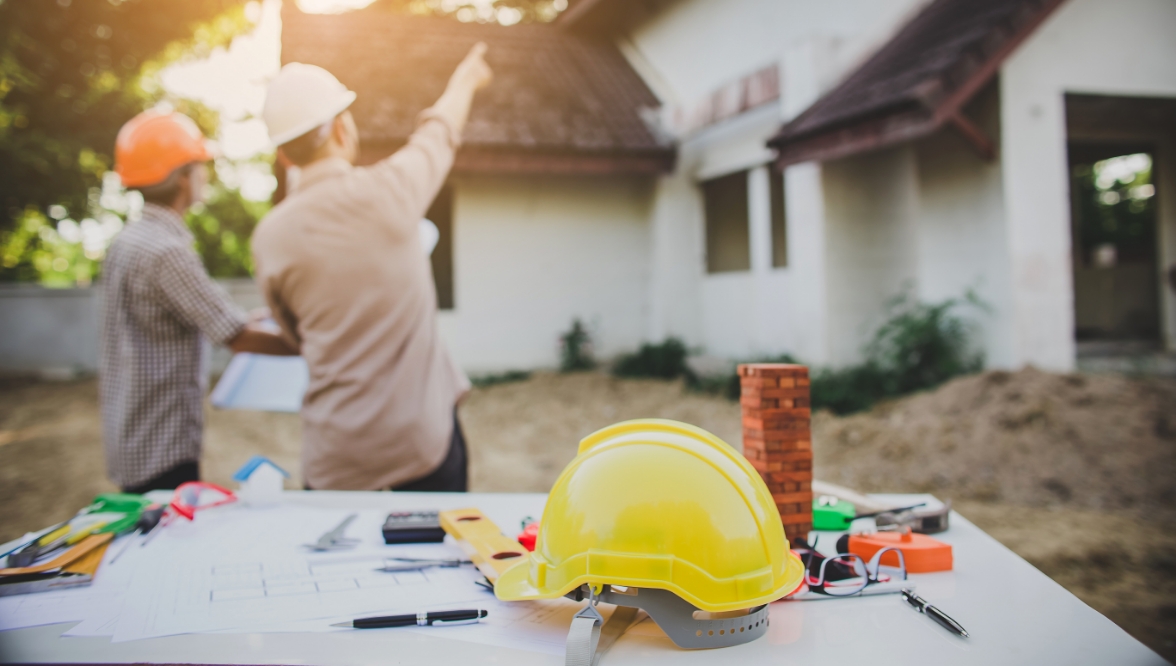 Two construction workers wearing hard hats discuss plans in front of a house under construction, with one pointing towards the building. A table in the foreground holds a yellow hard hat, blueprints, and various tools.