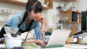 A young businesswomen wearing a blue shirt and black apron leans over a laptop smiling, while resting her elbow on the counter.