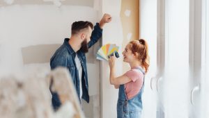 A young couple is standing in a partially renovated room, discussing paint colors while one person holds a fan of color swatches.