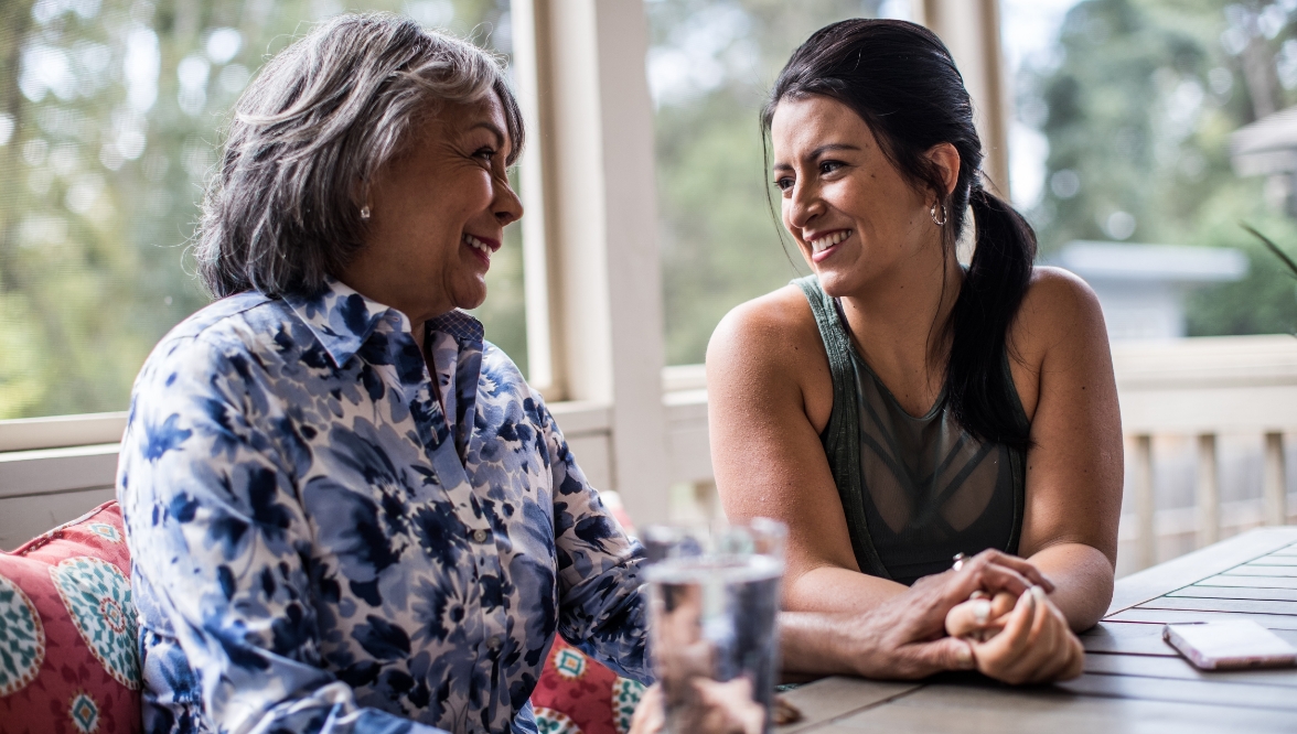 Two women, one older and one younger, are sitting together at a table on a porch. They are smiling warmly at each other, engaged in a friendly conversation, with a glass of water and a phone resting on the table.