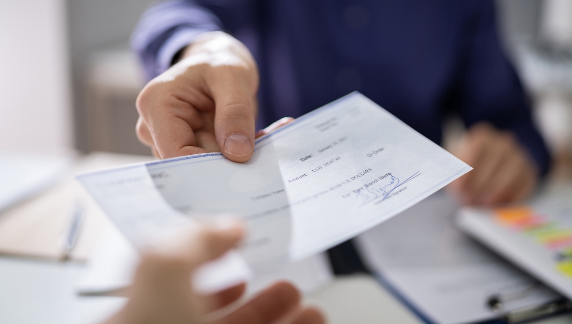 A close-up of someone in a dark blue shirt handing a check to someone else. Paperwork and folders are visible in the background