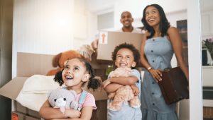 A joyful family stands in their new home, surrounded by moving boxes. Two young children, holding stuffed animals, look excitedly around while their smiling parents stand in the background with a box and suitcase.