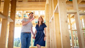 A man and women are holding hands and smiling while standing inside the wooden construction frame of a house.