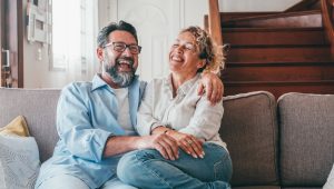 Couple sitting closely on a couch at home, sharing a moment of joy. Both are smiling and laughing while the man rests his hand next to hers.