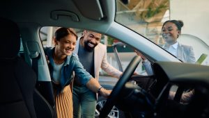 Image Alt Text A cheerful couple examines the interior of a new car, smiling as they interact with the car's dashboard, while a friendly salesperson stands nearby with a tablet, guiding them through the features.
