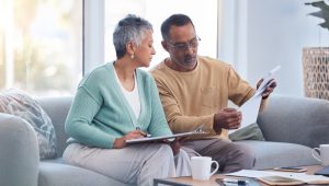 A mature couple sits together on a couch, reviewing paperwork with serious expressions. The man holds a document, while the woman holds a clipboard, appearing focused on the task at hand.