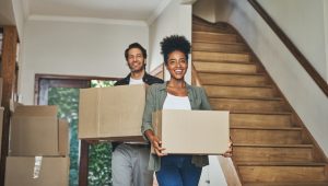 Two people carrying cardboard boxes in a house with a wooden staircase.