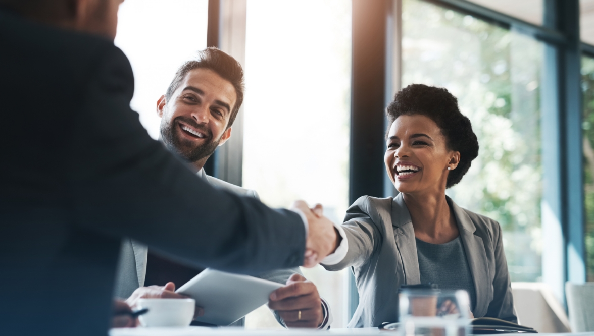 A group of professionals in a business meeting, with a woman and man smiling warmly as they shake hands across a table, symbolizing a successful partnership or agreement.