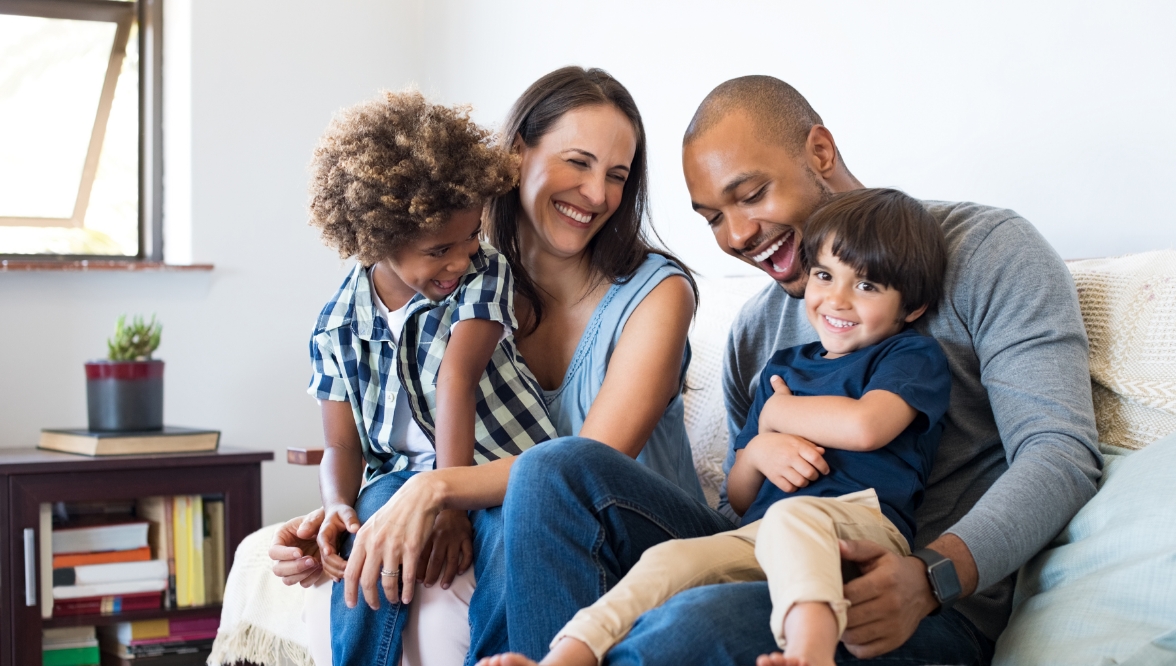 A family of two parents and two young children sitting on a couch in a living room, smiling and laughing. This image highlights care, togetherness, and the importance of planning for the children’s future.