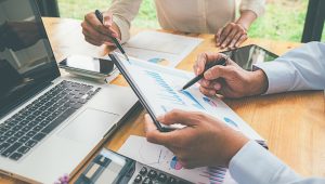 Two people are reviewing financial charts and graphs at a desk, holding pens and pointing to data on printed reports. The workspace includes a laptop, calculator, smartphone, and tablet, suggesting a business or financial planning discussion.