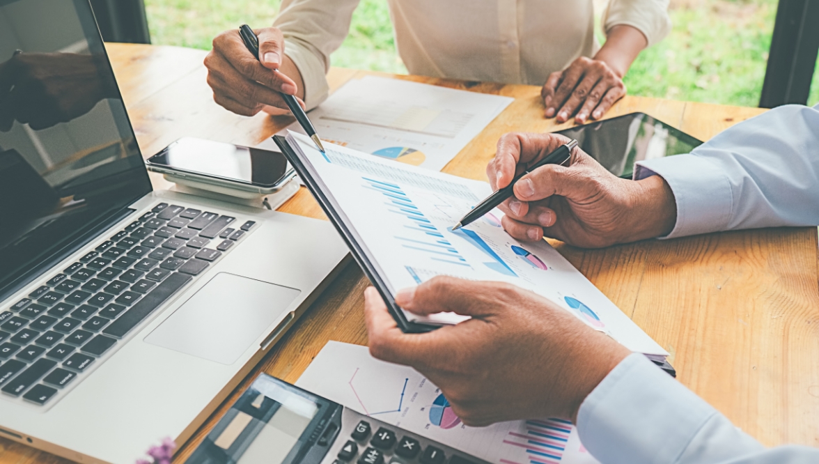 Two people are reviewing financial charts and graphs at a desk, holding pens and pointing to data on printed reports. The workspace includes a laptop, calculator, smartphone, and tablet, suggesting a business or financial planning discussion.