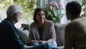 A concerned middle-aged woman sits on a couch with her hands clasped, listening intently to two other individuals, one older and one younger, in a warmly lit living room surrounded by lush greenery and flowers.