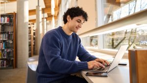 A male young adult sits on a stool at a countertop near a window in a library, working on his laptop. Rows of bookshelves can be seen in the background.