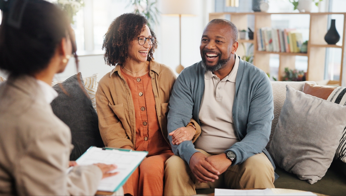 A cheerful couple sits on a couch smiling while engaging in conversation with a professional, who holds a clipboard and faces them in a relaxed, well-lit living room setting.