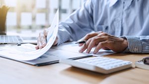A person wearing a striped shirt is sitting at a desk, reviewing documents and holding a pen. A calculator, laptop, and additional paperwork are visible on the desk.