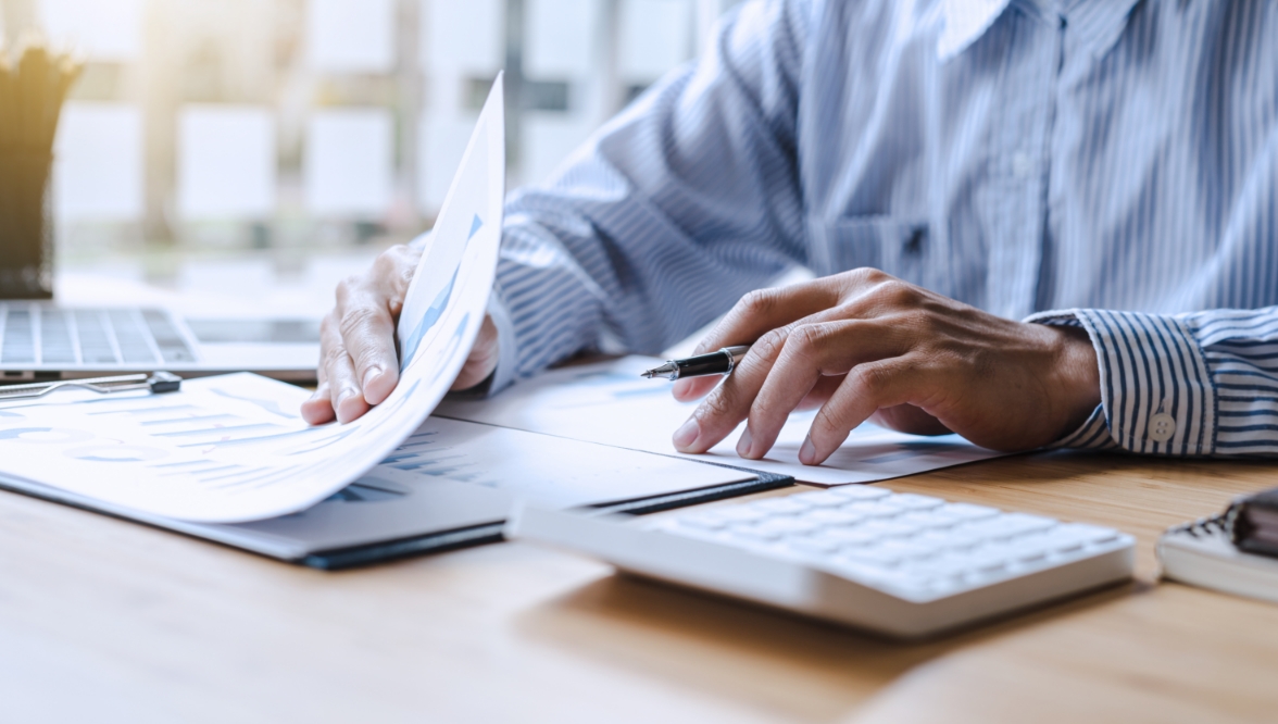 A person wearing a striped shirt is sitting at a desk, reviewing documents and holding a pen. A calculator, laptop, and additional paperwork are visible on the desk.