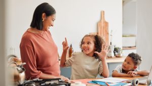 A cheerful mother smiles at her two young children at a kitchen counter. The older child excitedly raises her hands while the younger child looks on with curiosity. The counter is covered with school supplies, a laptop, and kitchen items.