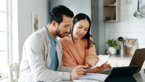 A couple sits in a brightly light kitchen reviewing paperwork together, a laptop and additional papers can be seen on the counter with them.