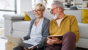 Happy senior couple at home sitting on the floor with a laptop and tablet.