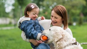 A smiling woman and young girl with Down syndrome are playing with a golden retriever holding an orange ball in a park.