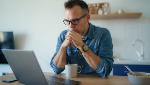 A thoughtful man sitting at a table with a laptop, appearing concerned while looking at the screen.