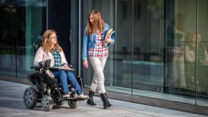 Two people outside a glass building, one in a wheelchair holding a laptop, the other carrying books.