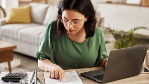 Person reviewing financial documents at a desk with a laptop and calculator in a bright living room setting.