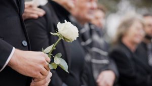 A person in a black suit holding a white rose, standing with others at a funeral.