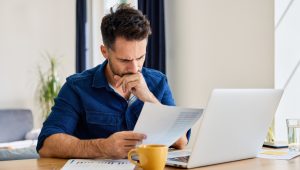 Person sitting at a desk, reviewing a financial document, next to an open laptop, coffee mug, and scattered papers.