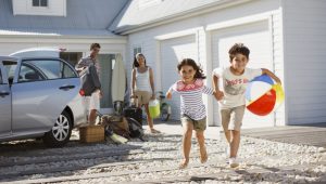 Family arriving at a vacation home, with two children running and parents unloading luggage from a car.