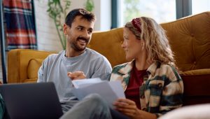 A happy couple sitting on the floor next to a sofa in a living room, discussing documents while using a laptop.