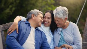 A smiling woman embraces her elderly parents while sitting on a wooden bench outdoors.