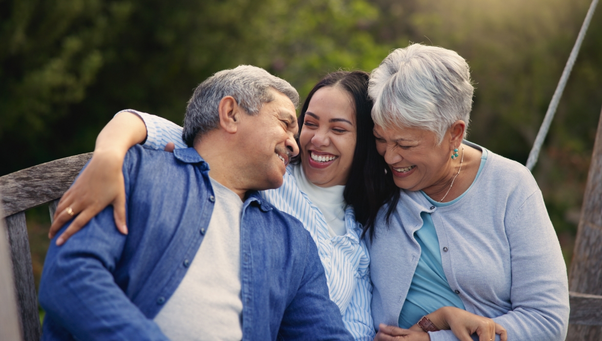 A smiling woman embraces her elderly parents while sitting on a wooden bench outdoors.
