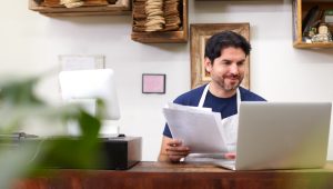 A man in an apron holds papers in his hand while referring to what’s on his laptop screen.