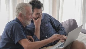 An older man reaches toward the screen of a laptop in a younger man’s lap.
