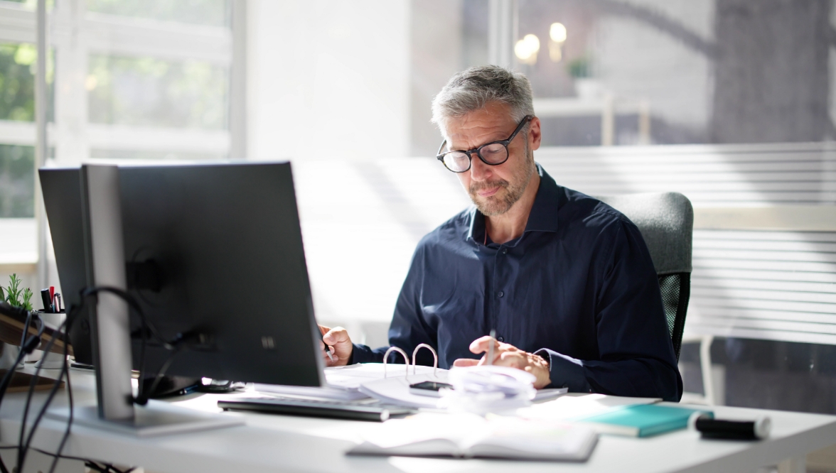 A man with gray hair works at a desk in an office, reviewing documents.