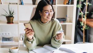 A smiling young woman sitting at a desk covered with notebooks and papers, holding a credit card in one hand and a smartphone in the other.