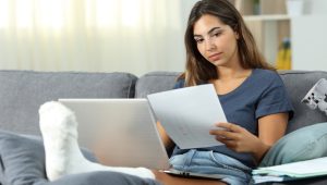 A young woman with her leg in a cast sits on a gray couch, focused on reading a document, with a laptop and additional papers nearby.