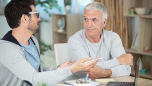 A younger man wearing glasses, a hoodie, and a T-shirt, explains something to a skeptical older man.