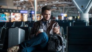 Sitting in an airport, holding a coffee, and flanked by his luggage, a young man checks his smartphone.