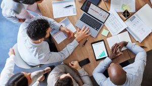 Five office workers, seen from above, examine financial data on a laptop and tablet and in various printed materials.