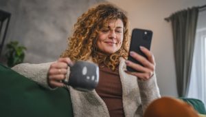 A woman with curly brown hair, a chenille sweater, and a beverage in a mug consults her smartphone.