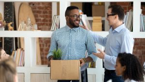 A man, wearing a blue shirt and glasses, holds a box containing a plant and smiles at a coworker who is happily wishing him well.