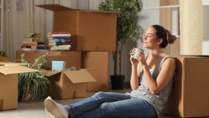 A young woman, leaning against a packing box with a mug in her hands, basks in the sunshine and her own independence.