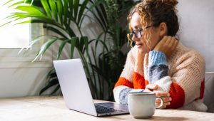 A woman wearing glasses and a striped sweater sits at a table with a laptop and coffee mug.