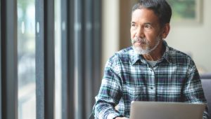 A bearded man wearing a plaid shirt looks up from his laptop and stares out the window.