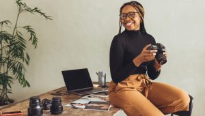 A photographer, perched on the edge of her desk, holds her SLR camera and smiles. Near her are photographs, lenses, and a laptop.